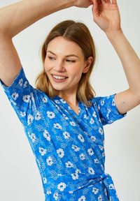 Blå blommig omslagsklänning med korta ärmar, prydd med vita blommor i olika storlekar och en midjeband för justerbar passform. Slät tygkvalitet.