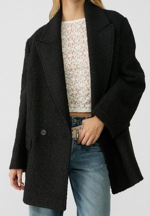 Kvinde iført en sort tekstureret oversized frakke over en hvid blonde crop top og blå jeans, justerer en beige bæltespænde.