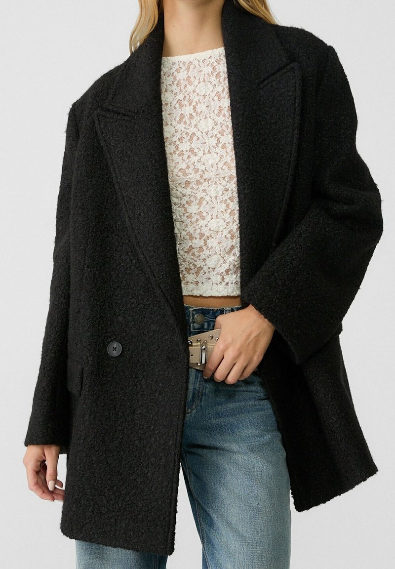 Femme portant un manteau oversize texturé noir par-dessus un crop top en dentelle blanche et un jean bleu, ajustant une boucle de ceinture beige.