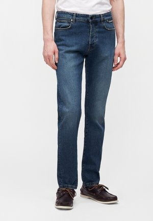 Man draagt donkerblauwe rechte jeans, wit overhemd ingestopt en donkerbruine leren veterschoenen, staand tegen een effen achtergrond.