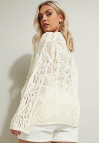 Off-white stickad tröja med ett geometriskt öppet vävmönster, avslappnad passform och sänkta axlar, bärs över vita shorts.