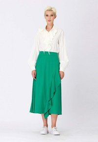 Blouse blanche à volants et jupe midi plissée verte avec un volant sur le côté, assorties à des baskets blanches. Lignes simples et épurées avec une couleur vibrante.
