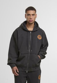 Sötét szürke cipzáras kapucnis pulóver kenguruzsebbel és hímzett logóval a bal mellrészen. Texturált szegéllyel és széles ujjal rendelkezik.