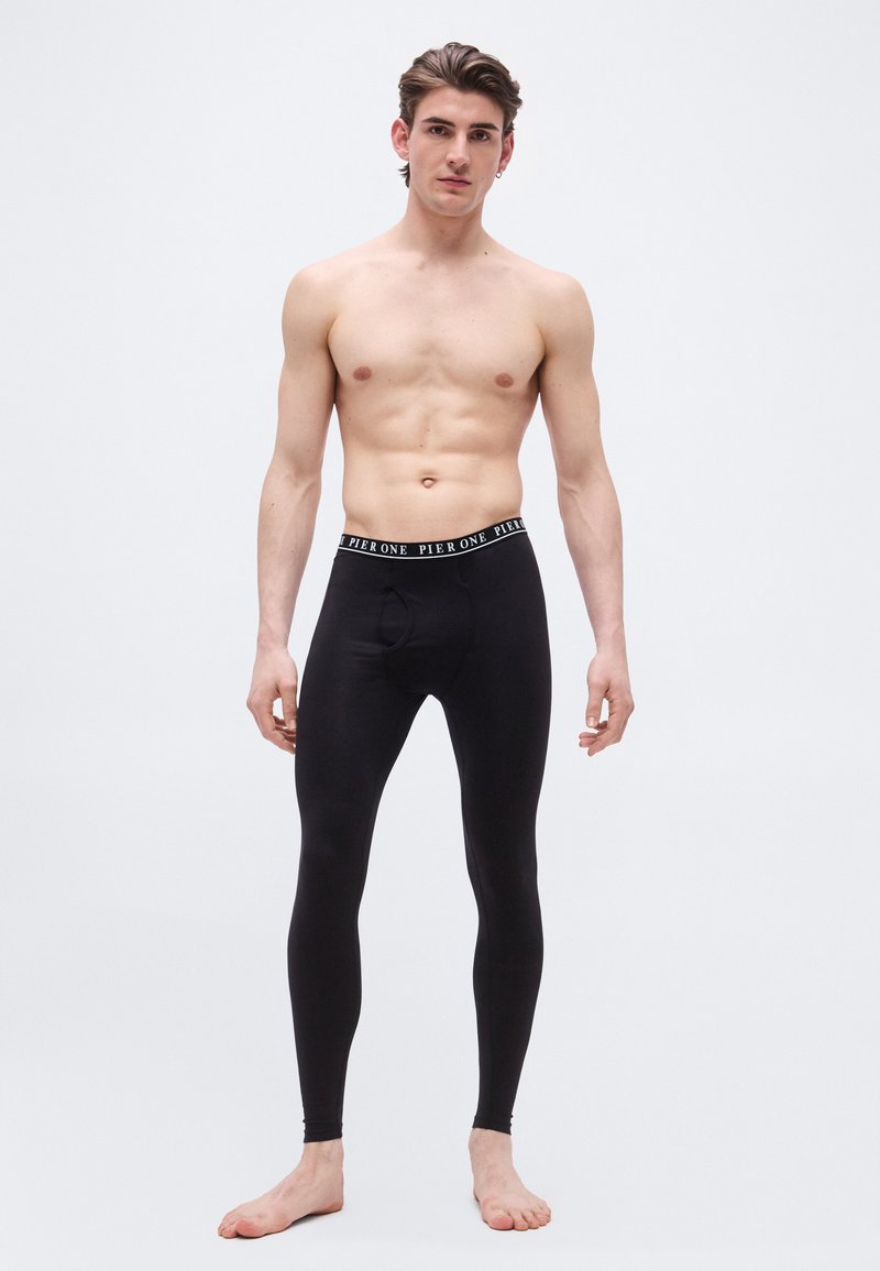 Jeune homme debout pieds nus portant un caleçon long noir avec la ceinture marquée "Pier One" sur un fond blanc uni.