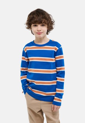 Enfant aux cheveux bouclés bruns portant un haut bleu à manches longues avec des rayures horizontales beige et blanches et un pantalon beige, debout avec une main dans la poche.