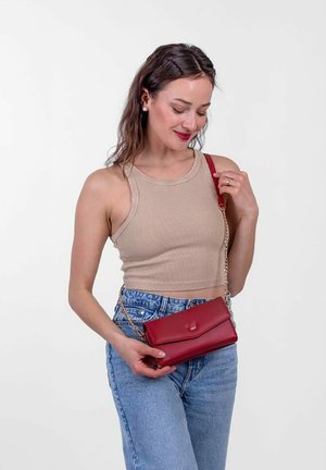 Červená kožená crossbody taška s řetízkem, skládací design a malý emblém vpředu, předvedená osobou v neformálním oblečení.