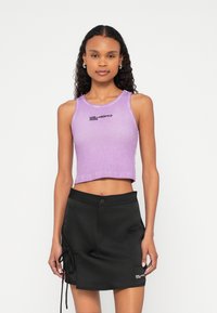 Mladá žena s kučeravými vlasmi nosí fialový krátky nástenný top a čiernu mini sukňu so zaväzovaním na boku a predným vreckom na bielom pozadí.