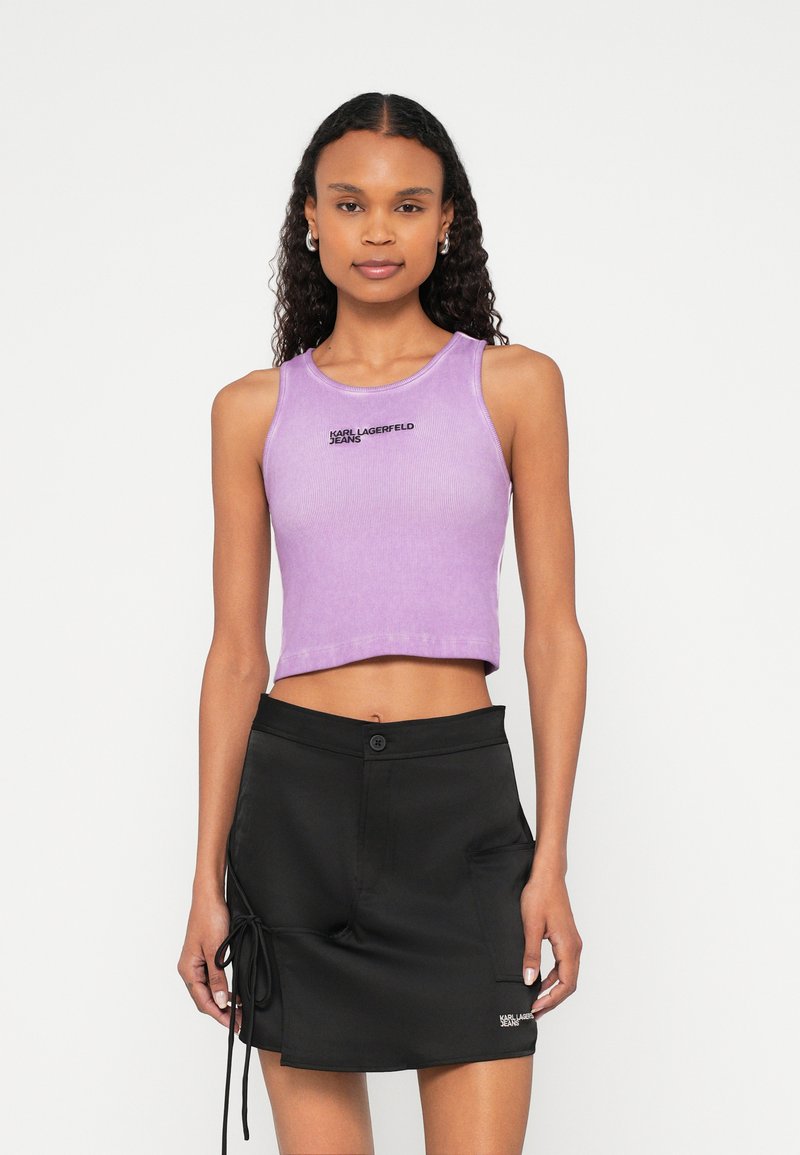 Mladá žena s kučeravými vlasmi nosí fialový krátky nástenný top a čiernu mini sukňu so zaväzovaním na boku a predným vreckom na bielom pozadí.