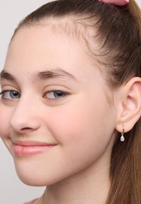 Złoty kolczyk w kształcie obręczy z małym, przezroczystym wisiorkiem w kształcie łzy, wiszącym na małżowinie usznej w zbliżeniu.