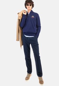 Navy zippered sweater over a light blue collared shirt, dark pants, and beige sneakers. Model holds a light tan coat; sunglasses on.
