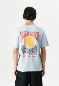 Lichtblauw t-shirt met korte mouwen met een kleurrijk grafisch ontwerp op de achterkant, met een zon, bergen en de tekst "REALISE FEED YOUR MIND."