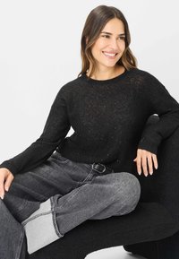 Schwarzer gestrickter Pullover mit glitzernden Akzenten, Rundhalsausschnitt und langen Ärmeln; getragen mit grauen, hoch taillierten Jeans mit aufgerollten Säumen.