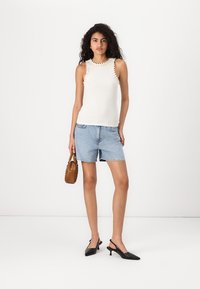 Camiseta sin mangas blanca con ribete decorativo, combinada con pantalones cortos de mezclilla azul claro. Tacones negros tipo slingback y un bolso marrón.