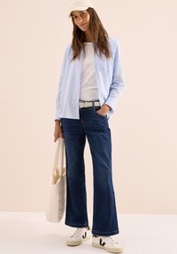 Ljusblå randig skjorta med knappar, vit t-shirt under, mörka denim jeans med vida ben, vit tote-väska och vita sneakers med svart logotyp.