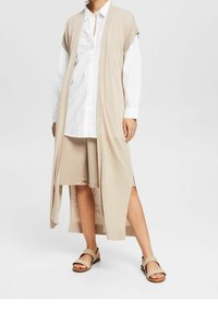 Beige lange Strickjacke mit offener Front, gestrickte Textur, kurze Ärmel über einem weißen Hemd mit Knöpfen, kombiniert mit einem beigen Rock und Sandalen.