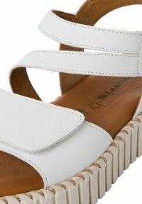 Sandale blanche avec des brides croisées, en cuir, et une semelle texturée beige. Comprend un footbed marron et des détails de couture visibles.
