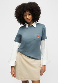 Giovane donna con capelli ricci che indossa una maglietta blu sopra una camicia bianca con colletto e una gonna beige, in piedi davanti a uno sfondo chiaro.