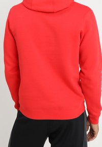 Röd huvtröja med långa ärmar som bärs av en person som visas bakifrån, ihop med svarta byxor.