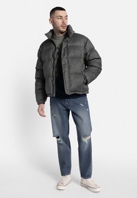 Párnázott, sötétszürke kabát magas gallérral, sötétzöld pulóverrel és koptatott kék farmernadrággal párosítva. Fehér tornacipő teszi teljessé a megjelenést.