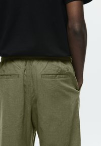 Olivgrüne Hose aus strukturiertem Stoff, mit einem Kordelzug an der Taille, zwei Gesäßtaschen und einem kleinen gestickten Logo auf der Rückseite.
