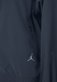Tumes sinine kangast jakk, millel on lukuga tasku, elastne alläär ja valge Jumpman logo vasakul poolel. Sile, kerge tekstuur.