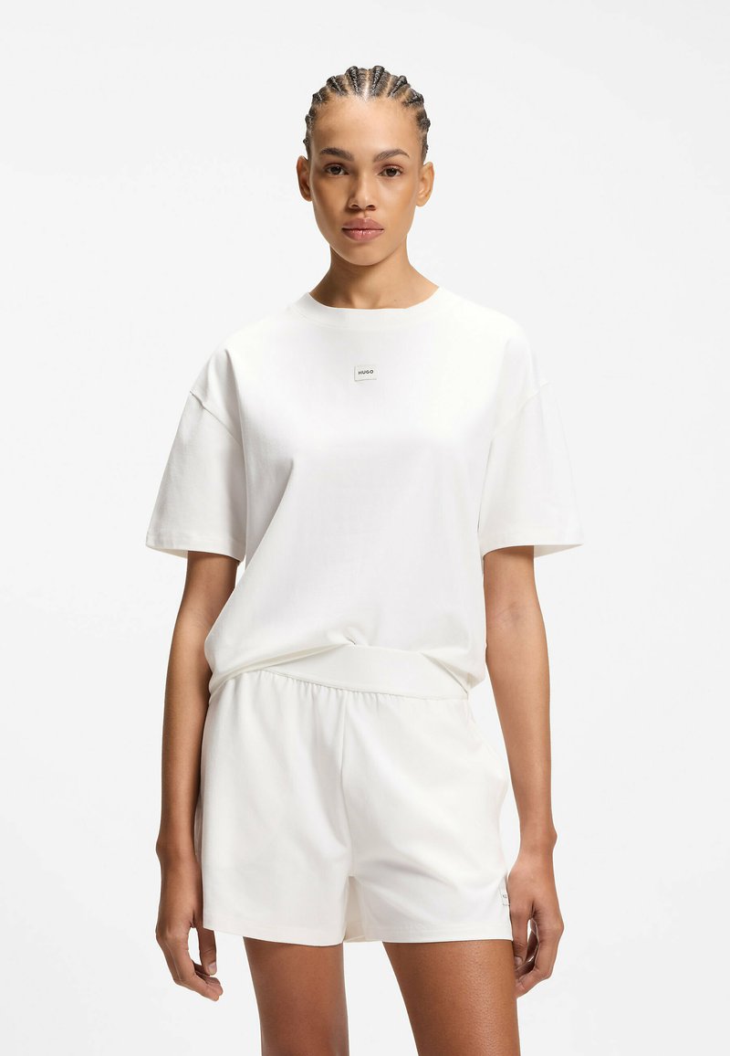 Femme aux cheveux tressés portant une tenue décontractée entièrement blanche, comprenant un t-shirt à manches courtes et un short assorti, debout sur un fond uni.
