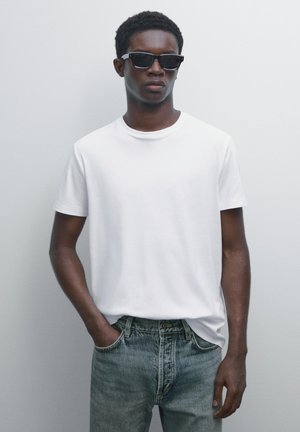 Junger Mann mit schwarzen Sonnenbrillen, weißem T-Shirt und blauen Jeans, steht mit der linken Hand in der Tasche vor einem schlichten Hintergrund.