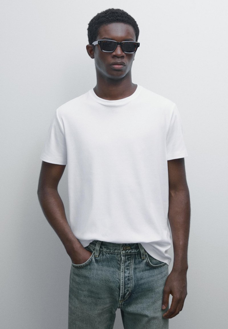 Ung mand iført sorte solbriller, hvid t-shirt og blå jeans, stående med venstre hånd i lommen mod en ensfarvet baggrund.