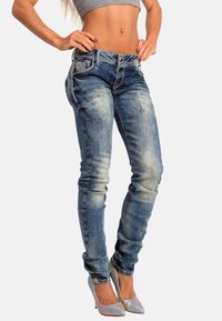 Jeans skinny in denim blu sbiadito con cuciture intricate, tasche decorate e leggermente usurati, abbinati a tacchi alti argentati scintillanti.