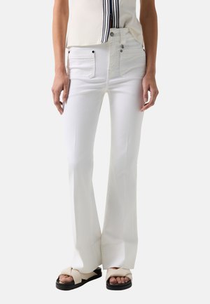 Kvinde iført hvide højtaljede bootcut-bukser, kort hvid top med sorte striber og beige sandaletter med krydset remme og plateausål.