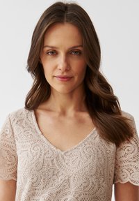 Béžová krajková blůza s krátkými rukávy, zdobená složitými květinovými vzory a dekoltem ve tvaru V. Měkká textura a jemné detaily po celém povrchu.