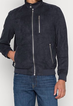 Homme portant une veste bomber en daim bleu marine foncé avec des fermetures éclair argentées et un jean bleu, main dans la poche gauche, debout devant un fond uni.