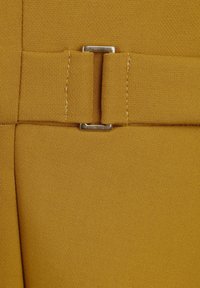 Gros plan d’un tissu jaune moutarde avec une boucle de ceinture cousue tenant une petite boucle métallique rectangulaire argentée.