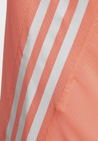 Camiseta atlética de color coral con tres franjas verticales blancas, hecha de un tejido texturizado. El dobladillo presenta costuras reforzadas a lo largo de los bordes.