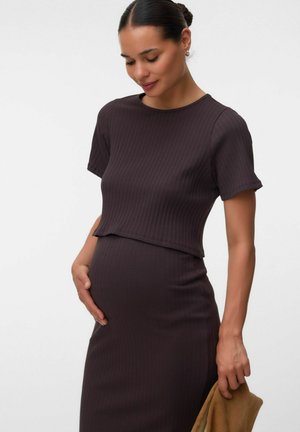Gravid kvinde iført tætsiddende ribstrikket mørkebrun kjole, der forsigtigt holder sin mave med den ene hånd og et foldet brun genstand i den anden.