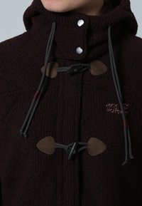 Burgunderfärgad stickad jacka med hög krage, med grå dragsko och bruna läderdetaljer. Logotypen är broderad i mörkröd.