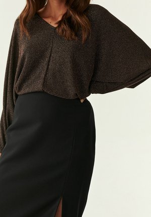 Femme portant un haut sombre à manches longues, ample, avec des rayures subtiles, et une jupe noire taille haute avec une fente à l'avant.