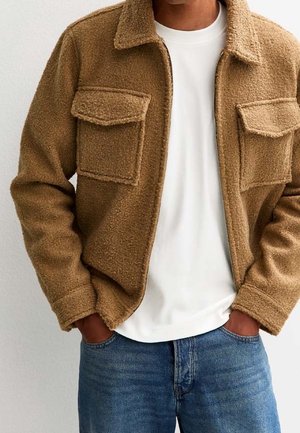 Man met een beige textuur ritsjack over een wit shirt, met handen in de zakken van een blauwe spijkerbroek, tegen een effen lichte achtergrond.