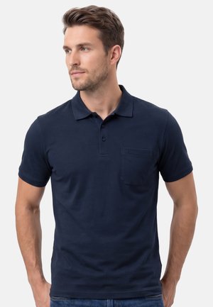 Mann trägt ein dunkelblaues, kurzärmeliges Polohemd mit Kragen und Brusttasche, schaut seitlich vor einem einfarbigen hellen Hintergrund.