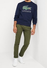 Marinblå sweatshirt med en grön krokodil-logotyp och vit text. Olivgrön byxa med avsmalnande passform. Vita sneakers fulländar outfiten.