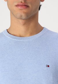 Lys blå strikkegenser med et teksturert mønster og en liten brodert logo i rød, hvit og blå på brystet.