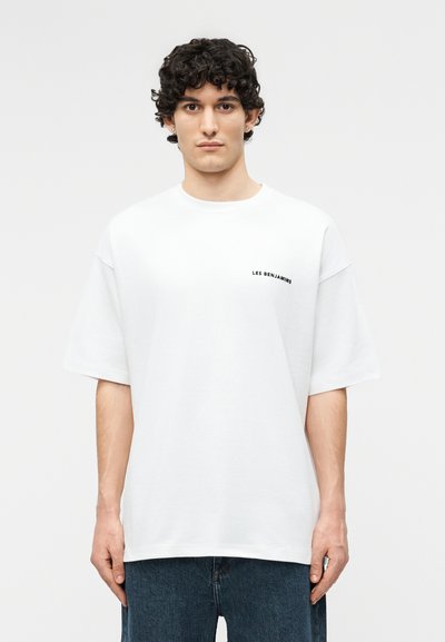Mladý muž s kučeravými tmavými vlasmi nosí voľné biele tričko s malým nápisom "LES BENJAMINS" na ľavej strane hrudníka a modré džínsy.