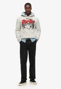 Felpa grigia con stampa del logo, indossata sopra una camicia azzurro chiaro. Pantaloni neri e sneakers completano il look. Stile casual e vestibilità rilassata.