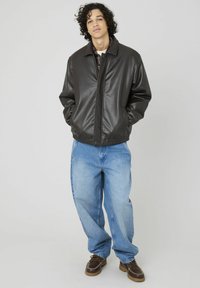 Veste en simili cuir noire avec col montant, fermeture à boutons-pression avant et ourlet élastique, associée à un jean baggy bleu clair et des chaussures brunes.