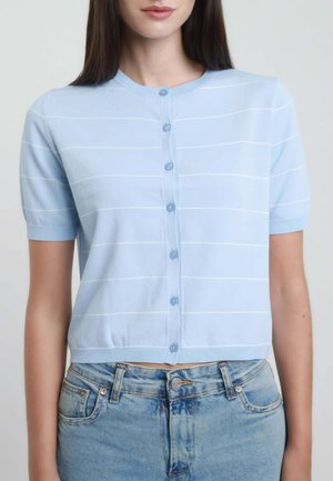 Donna che indossa un cardigan azzurro chiaro a maniche corte con bottoni, con sottili strisce orizzontali bianche e jeans azzurri.