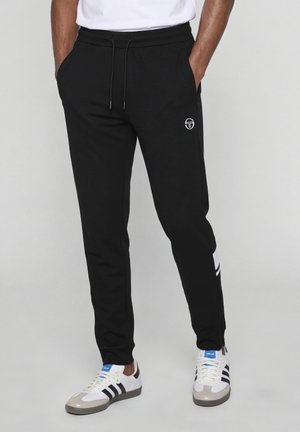Pantalon de jogging noir avec taille à cordon et petit logo blanc sur la cuisse, porté avec des baskets Adidas blanches à rayures noires.