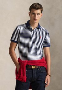 Young man wearing navy and white striped polo shirt, red sweater tied around waist, dark jeans, and brown leather watch.