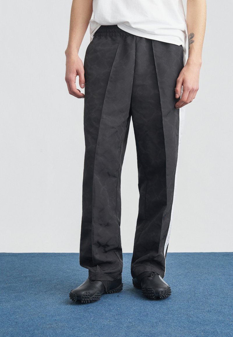 Άτομο που φοράει χαλαρά μαύρα παντελόνια με ρίγες στο πλάι και μαύρα υφασμάτινα παπούτσια, στέκεται σε μπλε χαλί μπροστά σε ανοιχτό γκρι φόντο.