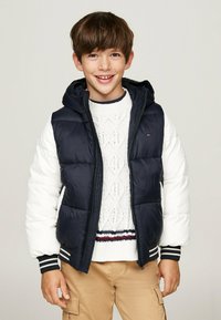 Giacca imbottita con un design a blocchi di colore blu navy e bianco, dotata di cappuccio, chiusura con zip e polsini a righe. Indossata sopra un maglione texturizzato.
