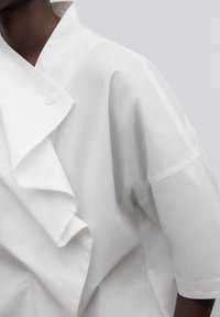 Camicia bianca in cotone con colletto strutturato e dettaglio a volant, maniche corte e chiusura a bottoni al collo.